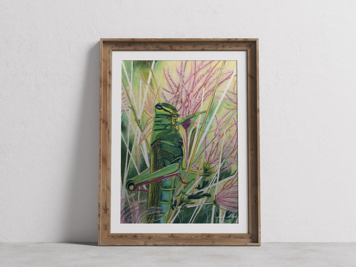 A giclee print of a bright green grasshopper in muhly pink grass in a wood frame with a white mat.