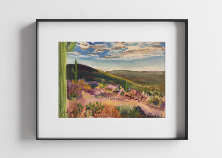 A giclee print of a pretty desert vista painting in a black frame with a white mat.