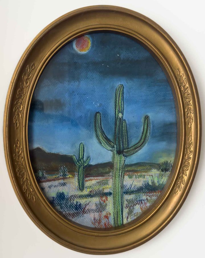 A vintage gold oval frame holds a print that depicts two cactus, an orange red moon and a few stars in the desert night.