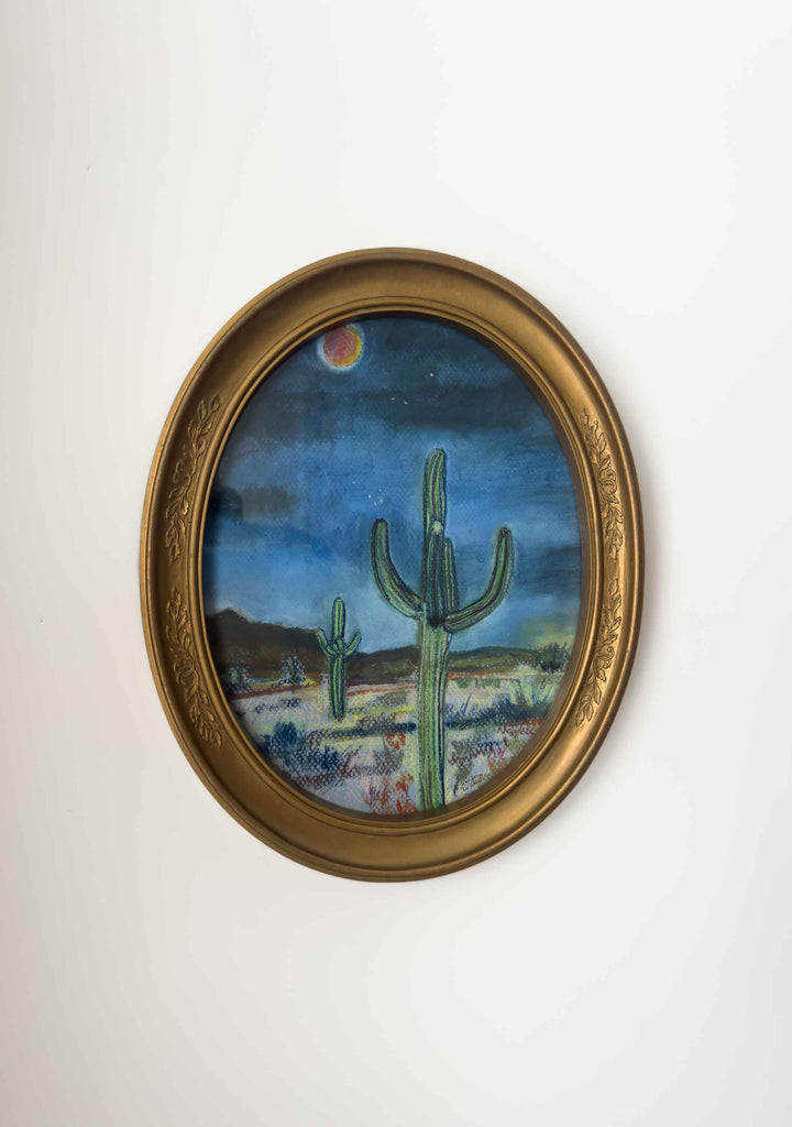 A pretty vintage gold oval frame holds a print of the desert at night. This print depicts two cactus and an orange red moon over the desert outside of town.