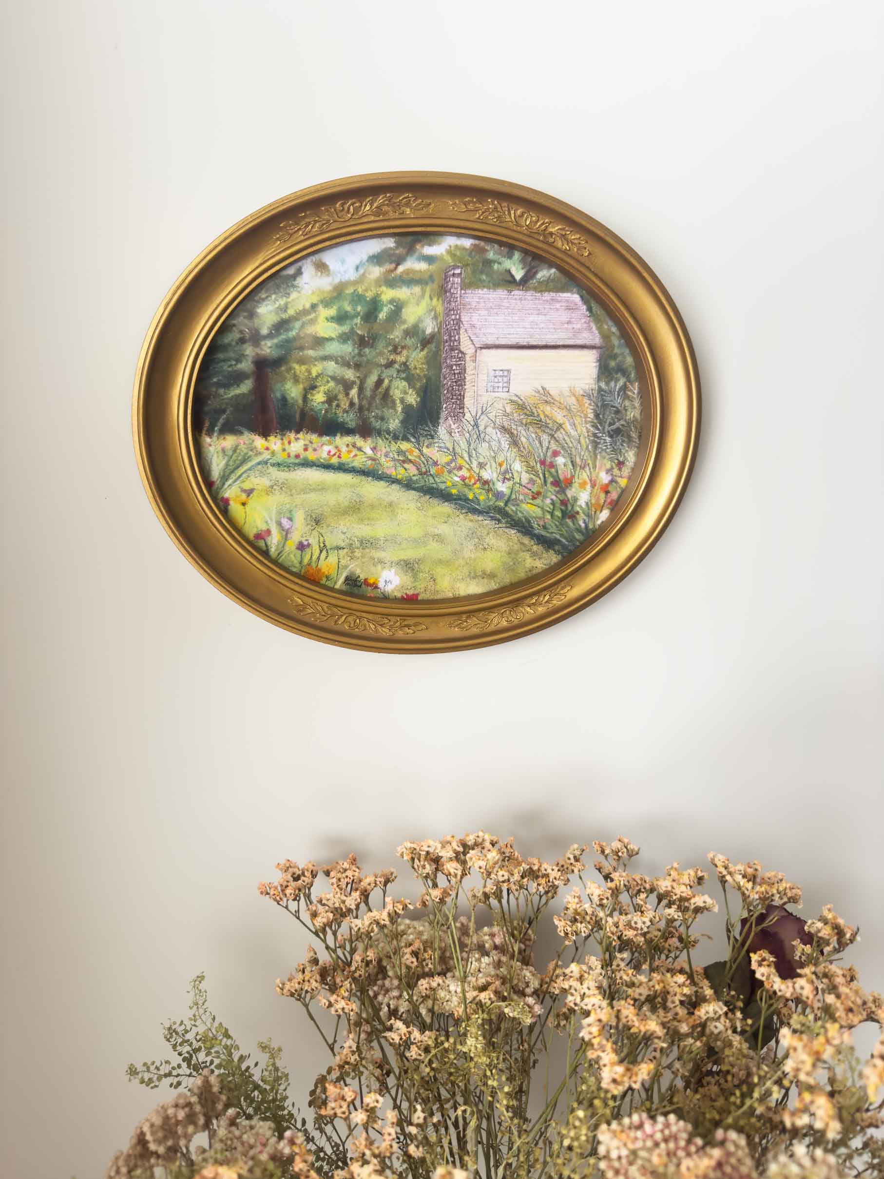 A flower garden and home at Shaker Village are displayed in the vintage, gold, oval frame.