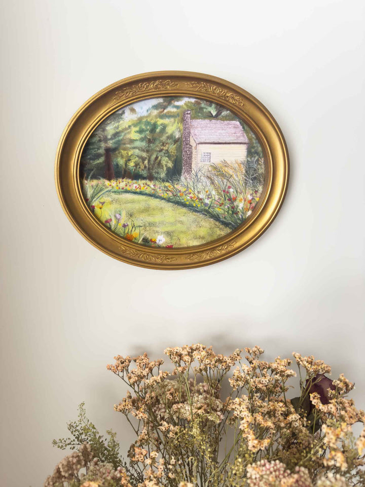 A flower garden and home at Shaker Village are displayed in the vintage, gold, oval frame.