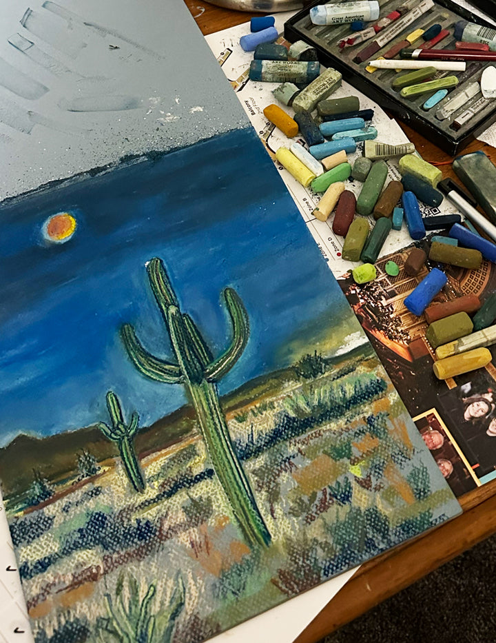 Soft pastels next to the painting of two cactus at night in the desert with the red moon above.