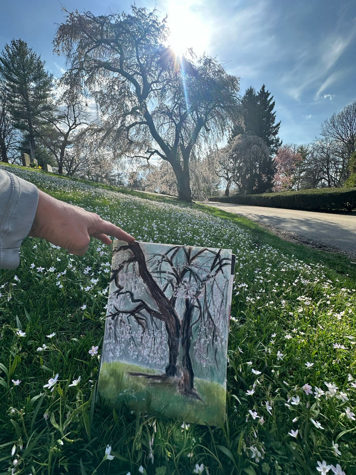 Artist Jennifer Millard hold up her plein air piece in front of the tree she is painting as she paints a weeping cherry blossom tree at the Lexington, Kentucky cemetery.
