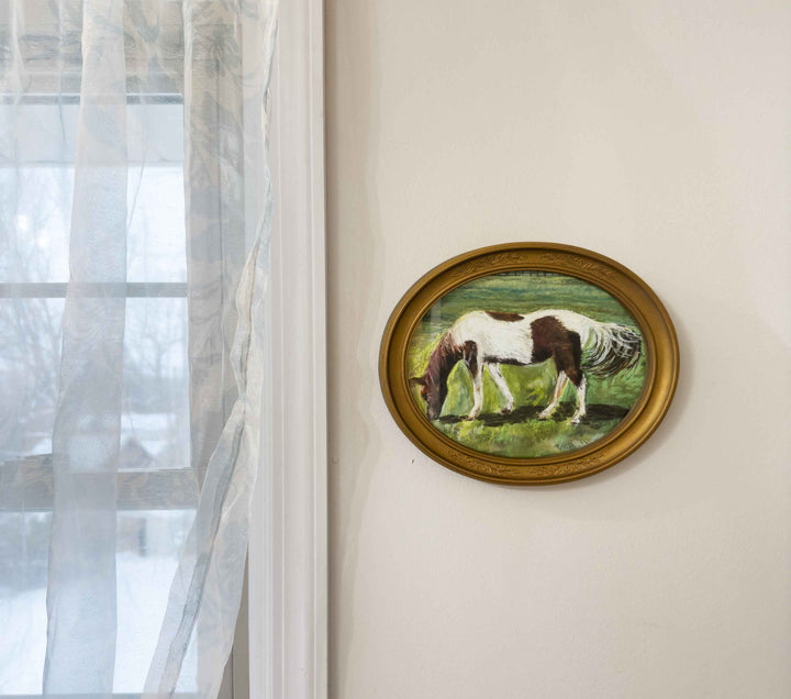 A photo of a print of a horse painting that used soft pastel displayed a vintage gold oval frame near a window.